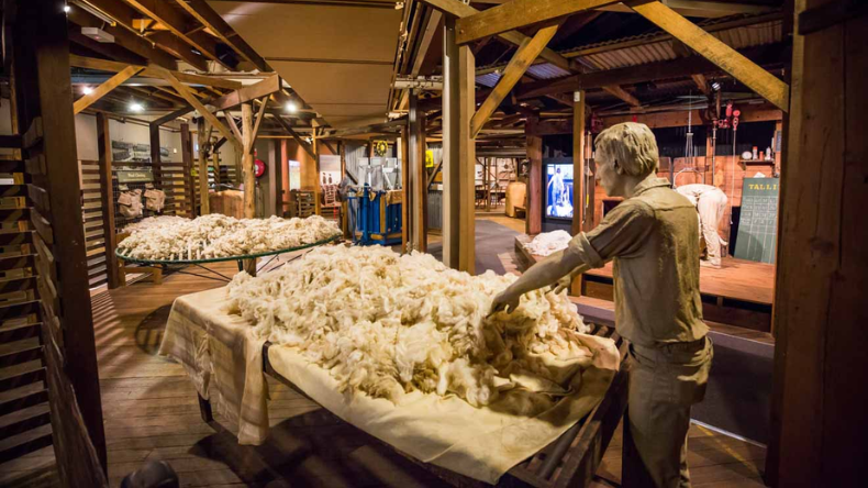Museum exhibit showing raw wool on large tables, a wool-sorting figure, and historic timber structures in a heritage industrial setting.