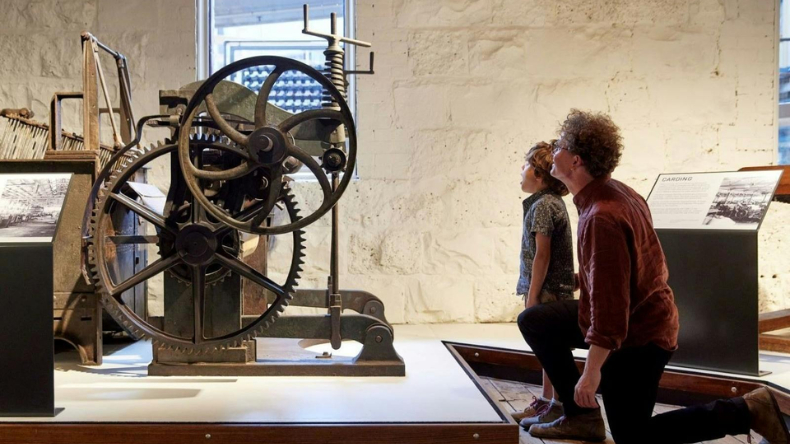 Two visitors viewing a large historic machine with exposed gears and wheels in a museum exhibit.