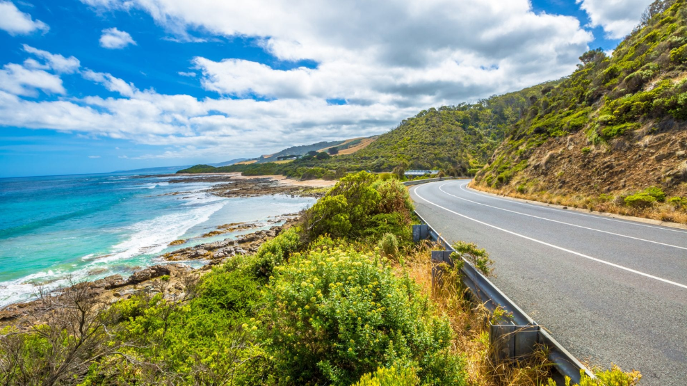 Coastal great ocean road curving beside turquoise ocean water and green hills under a blue sky with scattered clouds.