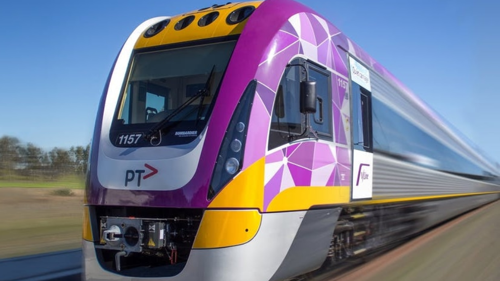 V/Line Modern purple, yellow and silver passenger train travelling at speed on a railway line under a clear blue sky.