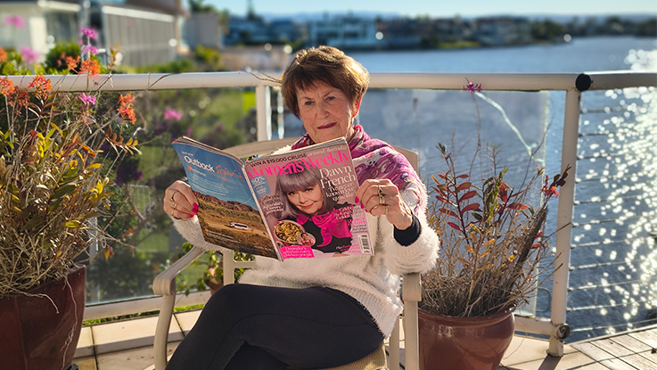 A woman sits on a waterfront balcony reading an Australian Women's Weekly magazine while relaxing in the afternoon sun. She wears a white knit jumper and a pink patterned scarf, with potted plants on either side of her chair. The calm water and distant shoreline create a bright, scenic backdrop.