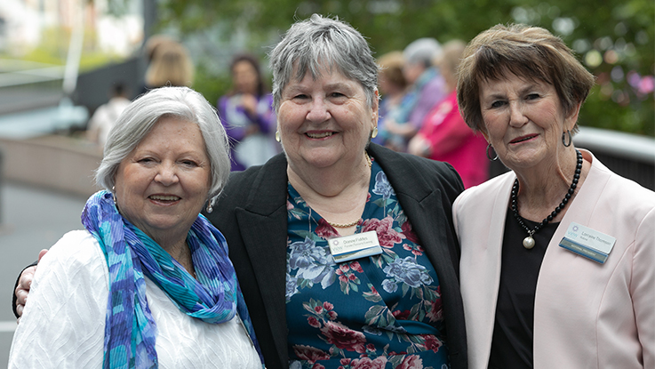 Three smiling women stand arm in arm outdoors at a community event, looking at the camera. The woman on the left wears a white top with a blue and turquoise scarf, the woman in the centre wears a floral top with a black jacket, and the woman on the right wears a pale pink blazer over a black top. All three wear name badges, with other attendees and greenery softly blurred in the background.