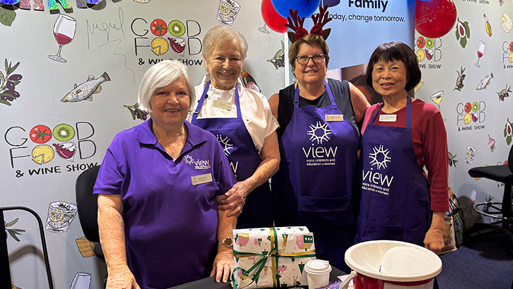 Four smiling volunteers wearing purple VIEW aprons pose behind a display table at the Good Food & Wine Show. The group stands in front of a branded event backdrop decorated with food and drink illustrations, with festive red and blue balloons overhead. Gift-wrapped items, a takeaway coffee cup, and raffle containers are arranged on the table in front of them.